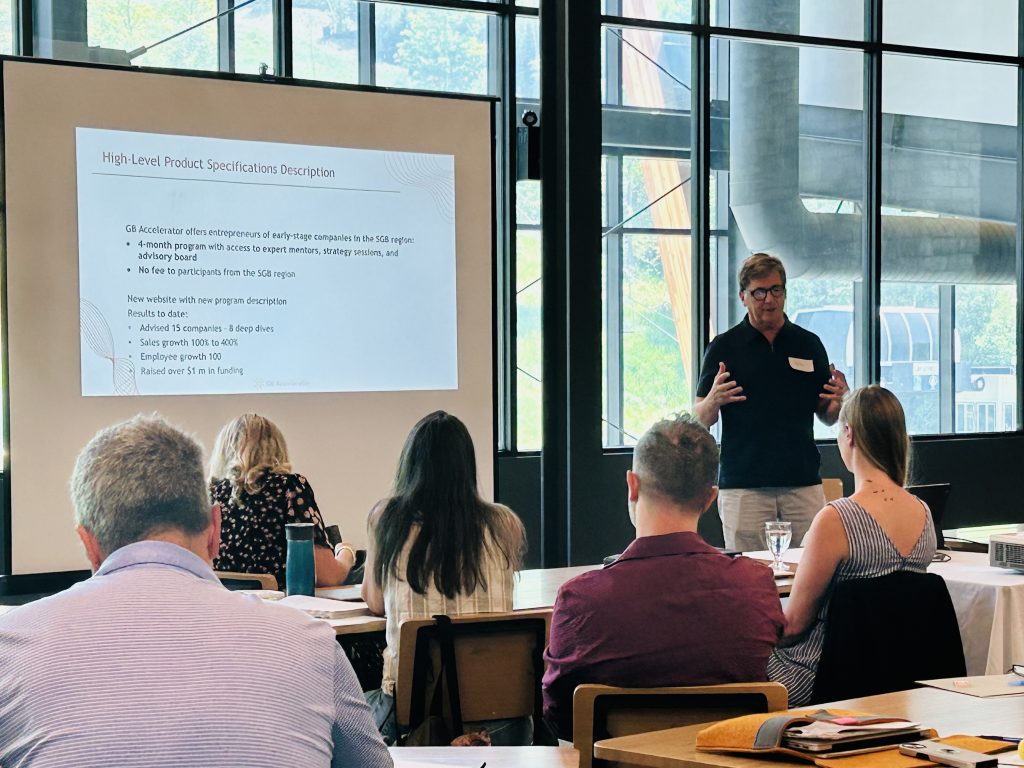 A speaker presents a slide to seated attendees in a bright conference room.