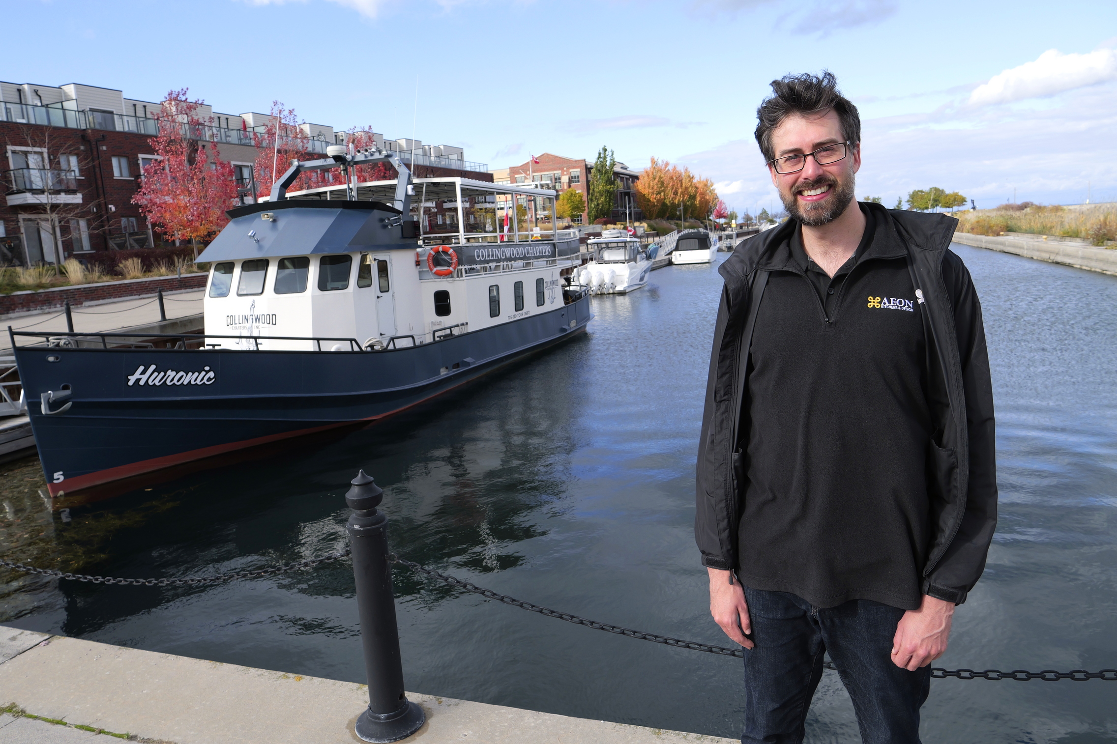 A man stands by a docked boat named Huronic on a calm canal with buildings in the background.