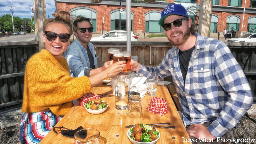 Three people sit at an outdoor table, toasting drinks with food in front of them on a sunny day.