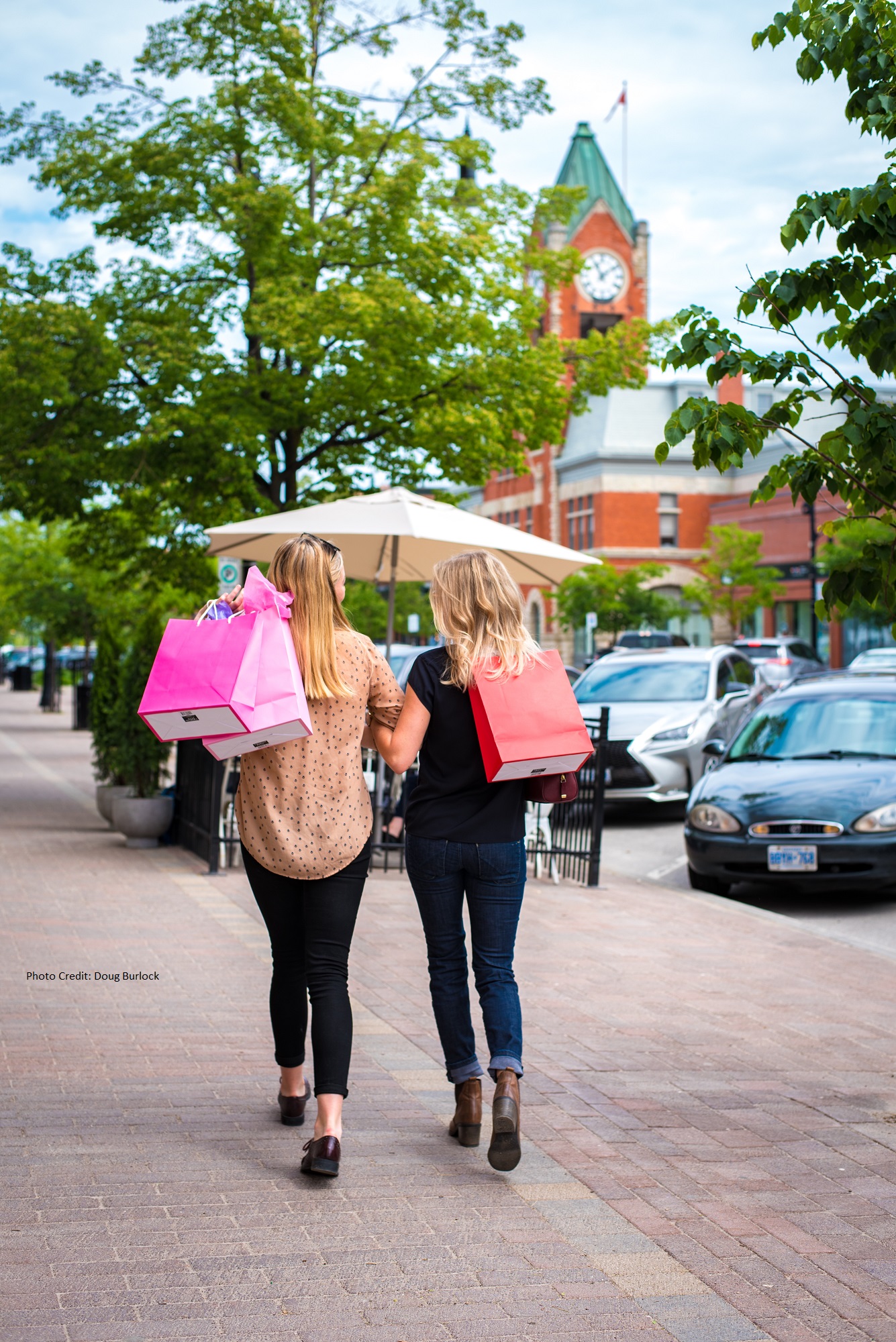 Two women walk down a sidewalk carrying shopping bags, with a clock tower in the background.