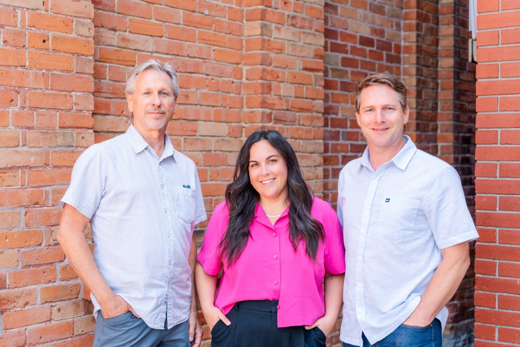 Three people stand in front of a brick wall, posing and smiling for the camera.